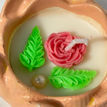 Decorative candle with pink rose, green leaves, and pearl in a terracotta holder.